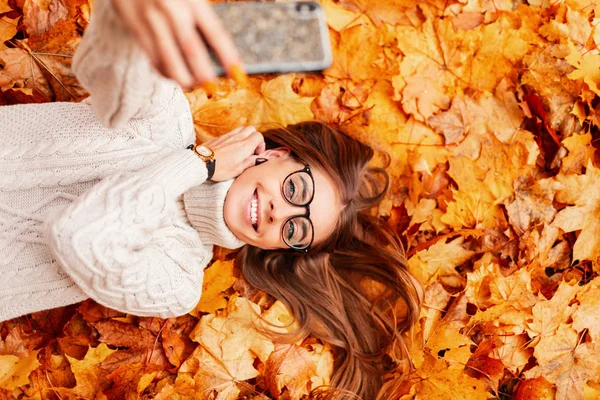 Приваблива щаслива молода хіпстерка в стильних окулярах в модному в'язаному светрі лежить на апельсиновому листі в парку і робить селфі по телефону. Красива дівчина посміхається. Вид зверху . — стокове фото