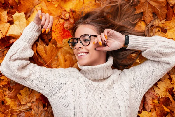 Belo modelo de mulher jovem feliz em um suéter tricotado em copos na moda relaxa deitado no parque na folhagem de outono dourada. Menina feliz atraente hipster descansando na floresta. Vista de cima . — Fotografia de Stock