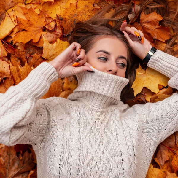 Moderna e elegante jovem com belos olhos azuis com cabelo castanho cobre o rosto com uma camisola vintage no fundo das folhas de outono. Menina bonito está descansando em folhagem laranja em um parque . — Fotografia de Stock