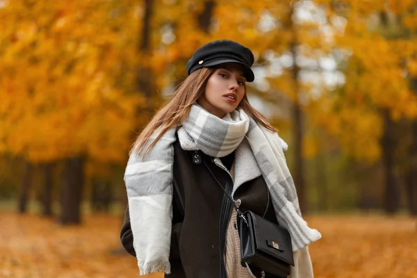 Sexy mooie jonge vrouw met een stijlvolle hoed in een bruine jas met een leren tas met een sjaal poserend op de achtergrond van bomen met oranje bladeren in het park op een herfstdag. Aantrekkelijk meisje buiten. — Stockfoto