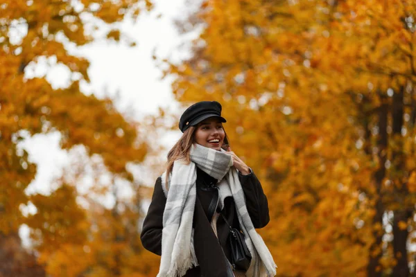 Trendy positive young woman model in fashionable warm clothes in a vintage hat with a scarf enjoys a walk in the park on a October day. Funny girl with a beautiful smile in the forest. Autumn fashion. — Stock Photo, Image