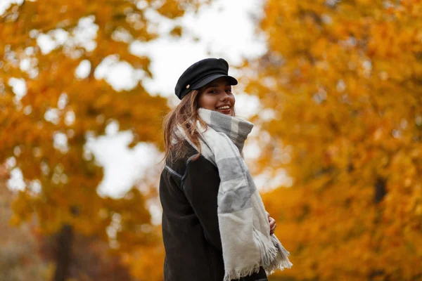 Jovem muito feliz em um belo sorriso em um chapéu na moda em uma jaqueta com um cachecol vintage posa em um parque em um fundo de folhagem laranja-amarela. Engraçado atraente elegante menina modelo ao ar livre . — Fotografia de Stock