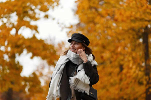 A mulher jovem na moda moderna em um chapéu em uma jaqueta com um lenço com uma bolsa feminina viaja no parque e positivamente sorri. modelo menina alegre caminha através da floresta e gosta da paisagem de outono . — Fotografia de Stock