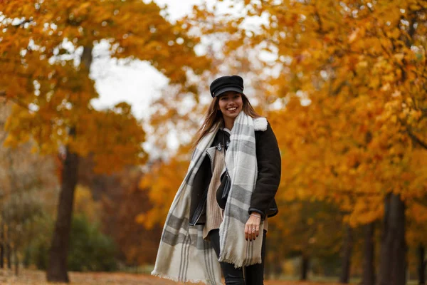 Carino allegra giovane donna in un cappello nero moda in una giacca vintage con una sciarpa calda viaggia attraverso il parco autunnale sullo sfondo di alberi con fogliame d'oro. Ragazza con sorriso positivo all'aperto — Foto Stock