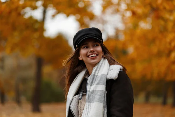 Modelo de uma jovem mulher positiva em um chapéu elegante em uma jaqueta marrom em um lenço para um passeio na floresta de outono. Menina alegre com um sorriso bonito está descansando no parque em um fundo de folhagem dourada . — Fotografia de Stock