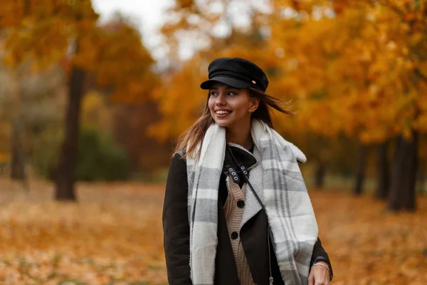 Jovem muito positiva com um belo sorriso na moda roupas quentes em um chapéu vintage com um lenço goza de um passeio no parque em um dia de outubro. Modelo de menina engraçada na floresta. Estilo outono . — Fotografia de Stock