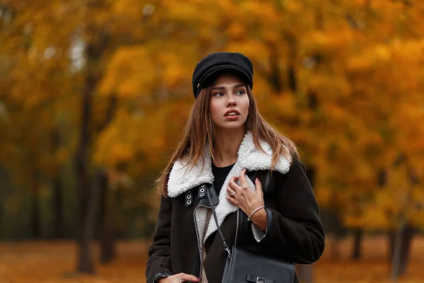 European stylish young woman fashion model in a trendy black hat with a leather bag posing in autumn park on a background of trees with orange foliage. Pretty elegant girl in the forest. Casual look. — Stock Photo, Image