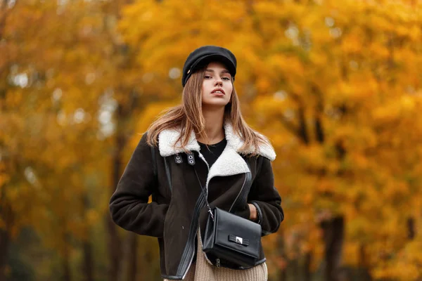 Mulher bonita bonita em uma jaqueta marrom elegante em um chapéu elegante com uma viseira com um saco de couro na moda posando no parque. Modelo de moda menina moderna em roupas de outono na moda ao ar livre . — Fotografia de Stock
