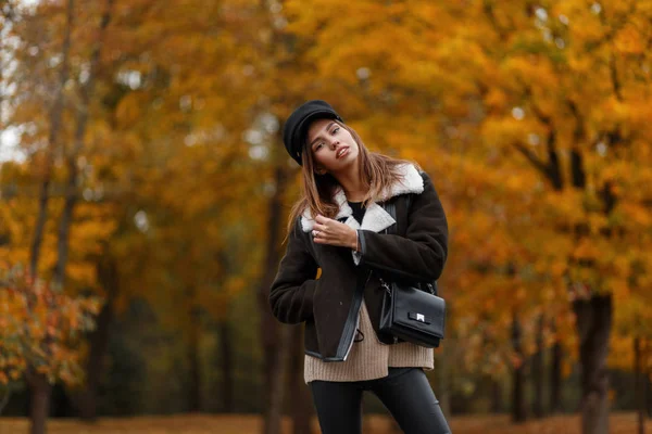 Pretty stylish model of a young woman in fashionable clothes in an elegant hat with a leather handbag posing in the park on an autumn warm day. Beautiful girl in trendy outerwear in the forest. Style. — Stock Photo, Image