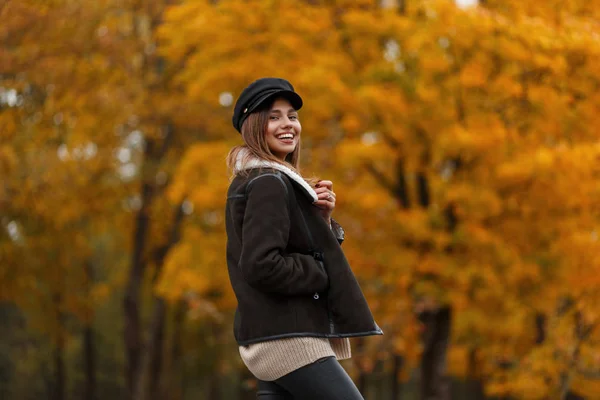 Jovem alegre com sorriso positivo em uma jaqueta em um chapéu na moda com um saco de couro posando no parque de outono em um fundo de árvores com folhagem laranja. Menina alegre na floresta. Olhar casual . — Fotografia de Stock