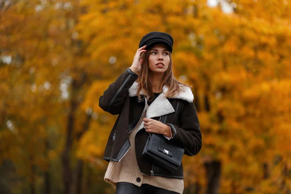 European young woman in stylish clothes in a vintage hat with a black leather bag posing in a park. Attractive trendy girl fashion model in the forest on a background of gold leaves. Autumn style. — Stock Photo, Image