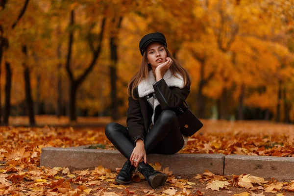 Stylowe cute młoda kobieta w modne ciepłe ubrania w skórzane modne buty w siada w parku na ziemi wśród pomarańczowych jesiennych liści. Nowoczesna piękna dziewczyna odpoczynek na świeżym powietrzu w lesie. — Zdjęcie stockowe