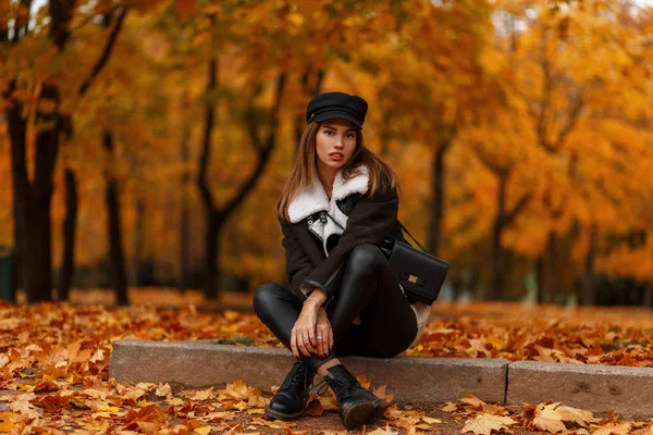 Modelo de uma jovem mulher elegante em uma jaqueta marrom vintage em botas pretas elegantes em um chapéu preto em leggings de couro na moda está descansando ao ar livre em um parque em um dia de outubro. Menina elegante atraente . — Fotografia de Stock