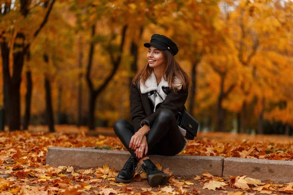 Pretty happy young woman in a chic hat in a stylish jacket with a bag relaxes in the forest on a background of golden leaves. Joyful girl sit in the park. Autumn collection of trendy women's clothing. — Stock Photo, Image