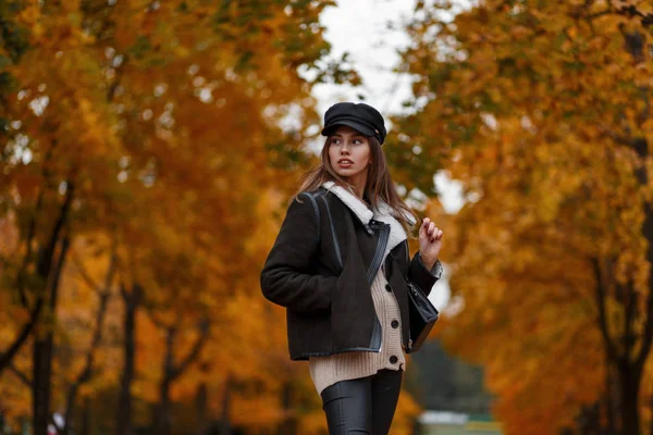 Linda modelo de moda de mujer joven con estilo en un sombrero negro de moda con una bolsa de cuero posando en el parque de otoño sobre un fondo de árboles con follaje naranja. Una chica muy elegante en el bosque. Aspecto casual . — Foto de Stock