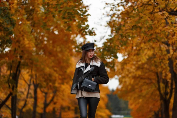 Jovem europeia moderna com roupas quentes e elegantes em um chapéu vintage com uma bolsa de couro preto viaja em um parque de outono. Modelo de moda menina muito na moda na floresta em um fundo de folhas de ouro . — Fotografia de Stock