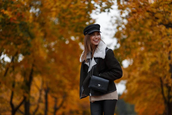 Joyful young woman in stylish outerwear in an elegant hat with a vintage bag stand in the park and enjoys the autumn scenery. Happy girl with a cute smile travels on the forest with yellow foliage. — Stock Photo, Image