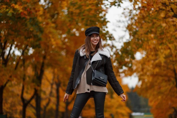 Élégante jeune femme heureuse dans un chapeau chic dans une veste élégante avec un sac dans la forêt sur un fond de feuilles d'or. Fille gaie marche dans le parc. Collection automne de vêtements femme tendance . — Photo