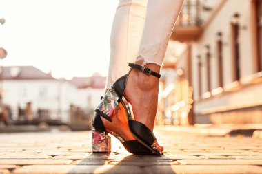 Beyaz kotlu, zarif deri sandaletli kadın bacakları gün batımında şehirde duruyor. Yakın plan..