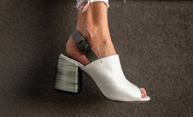 Gri arka planda kadın bacağında topuğu olan zarif, beyaz deri bir yaz ayakkabısı. 