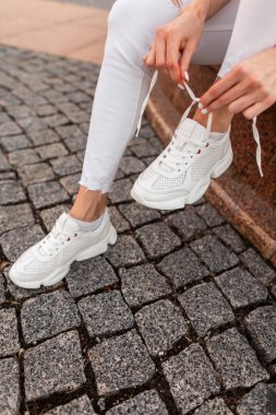 Şık kotlu kız oturur ve ayakkabı bağcıklarını sokaktaki beyaz moda ayakkabılarına bağlar.