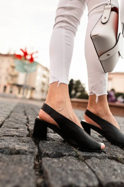 ファッショナブルな黒革の夏の靴で白いデニムパンツの女性の足のクローズアップは 通りにスタイリッシュなバッグ — ストック写真