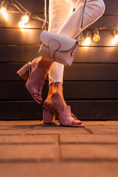 Kot elbiseli şık bir kadın yaz ayakkabılı bir çanta ile şehirde ahşap bir duvarın yanında poz veriyor..