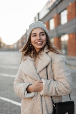 Eski moda şapkalı, modaya uygun siyah deri çantalı, sokakta tuğla binaların yakınındaki bir otoparkta poz veren ve gülümseyen modaya uygun genç bir kadın. Komik şirin kız modeli..