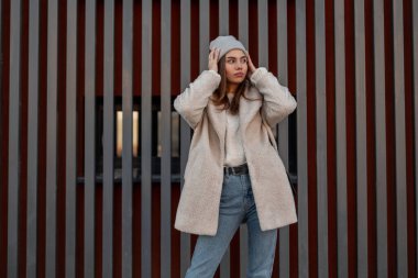 Kentin modern metal duvarının yanında, modaya uygun mavi renkli kot pantolonlu şık, genç bir kadın, modaya uygun gri örülmüş şapkasını düzeltiyor. Açık havada poz veren seksi manken kız..