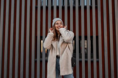 Modaya uygun örgü şapkalı, eko-kürk pozlarından sütün renginde komik, güzel bir genç kadın ve gri bir metal duvarın yanında gülümsüyor. Mutlu kız modeli. Bahar gençlik tarzı.