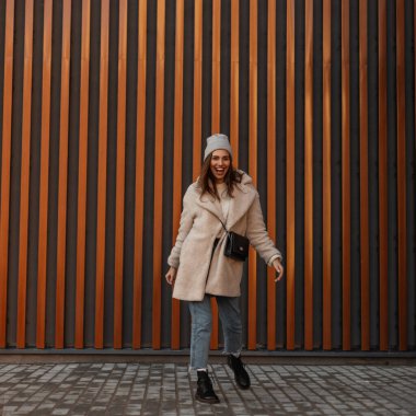 Kentin klasik metal duvarının yanında, deri çizmeli örülmüş bir şapkayla poz veren ve kahkahalar atan modern, oldukça olumlu ve modaya uygun genç kadın kıyafetleri. Mutlu kız manken ayakta duruyor ve gülümsüyor..