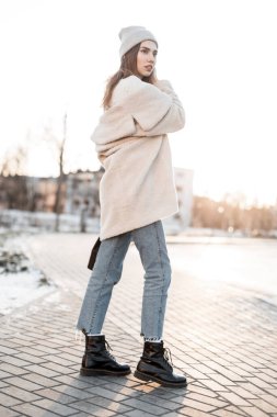 Parlak bir günde dışarıda poz veren modaya uygun siyah çizmeli, mavi kot pantolonlu şık bir eko ceketli, şık bir genç kadın. Moda mankeni yürüyüş ve kış güneşinin tadını çıkarıyor..