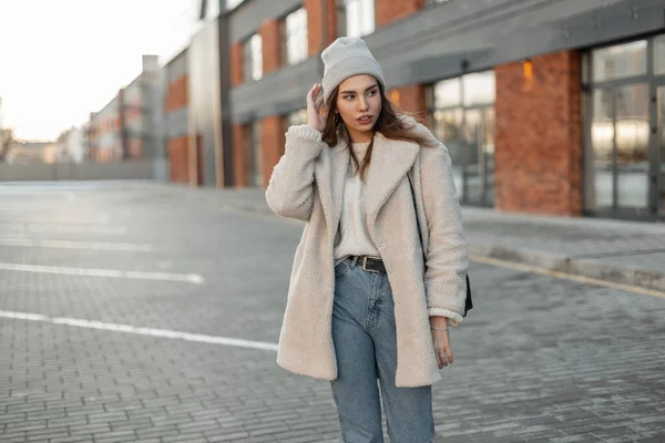 ジーンズのファッショナブルなフェイクファーコートを着たスタイリッシュなセーターのニット帽の魅力的な若い女性は レンガ造りの建物の近くの駐車場に立っています 市内の美しい女の子モデル屋外 — ストック写真