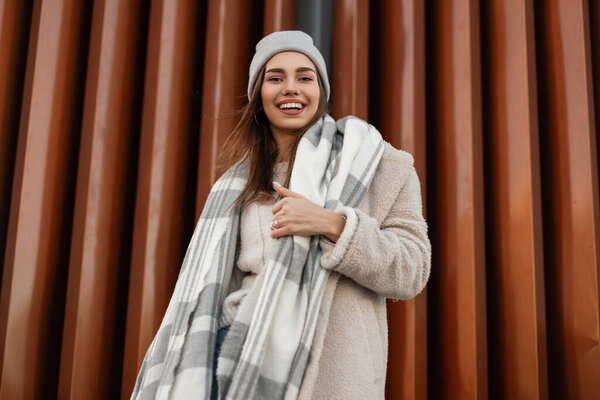Young positive woman in a fashionable knitted hat in a stylish coat of milk color from eco-fur with a woolen warm scarf poses and cute smiling near a vintage metal wall. Pretty funny girl model.