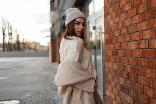 Beautiful young woman in a fashionable gray hat in a stylish knitted sweater in a youth spring eco fur coat posing near a vintage brick building on the street. Attractive girl model walks on the city.