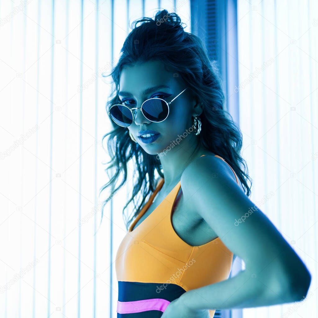 Stylish sexy young woman with curly hair in trendy sunglasses in a colored stylish swimsuit sunbathes in a solarium. Attractive hipster girl is standing in a modern tanning bed.