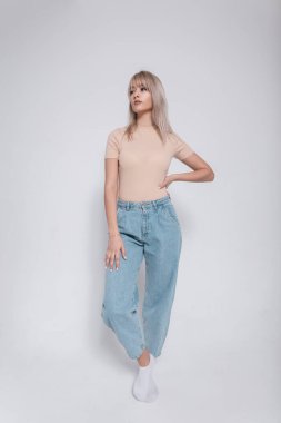 Modaya uygun kot pantolonlu, şık, genç, sarışın bir kadın içeride beyaz bir duvarın yanında duruyor. Stüdyoda gündelik giysiler içinde zarif modern kız model. Gençlik modern tarzı.