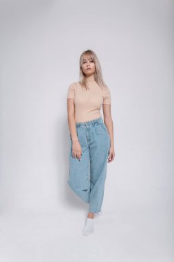 Moda mankeni, şık, mavi kotlu, modaya uygun bir gömlek giyen, beyaz bir duvarın yanında duran göz alıcı sarışın bir kadın. Güzel, zarif, genç ve şık bir kız.. 