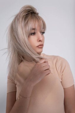 Beyaz arka planda, şık bej tişörtlü, sarışın bir kadının şık portresi. Çekici kız modeli içeride saç düzleştirir. Güzel bayan. Yakın plan..