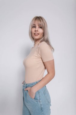 Oldukça neşeli genç bir kadının portresi. Pozitif bir gülümsemesi var. Odadaki beyaz bir duvarın arka planında sarı saçlar ve şık kıyafetler var. Odada poz veren neşeli model kız. Bahar stili