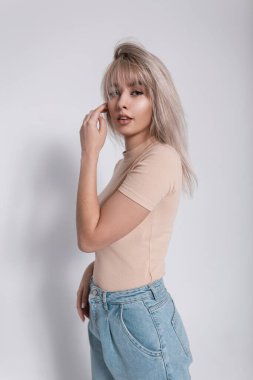 Moda mankeni, şık, mavi kotlu, modaya uygun bir gömlek giyen, beyaz bir duvarın yanında duran göz alıcı sarışın bir kadın. Modern, güzel, genç, zarif bir kız.. 