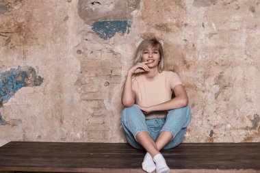 Havalı kotlu şık bir tişört giyen çekici sarışın bir kadının modaya uygun genç modeli odadaki eski bir tuğla duvarın yanında dinleniyor. İçeride gülümseyen sevimli mutlu kız modeli. Moda..