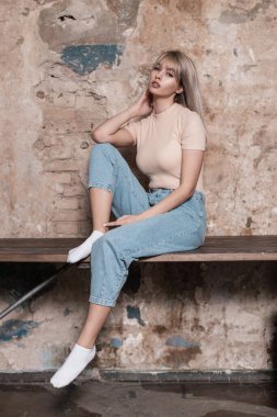 Şık kotlu, sarı saçlı, seksi dudaklı Avrupa modasına uygun genç bir kadın odadaki klasik tuğla bir duvarın yanında duruyor. Evde genç kız kıyafetleri içinde sevimli bir manken. Moda bakışı..