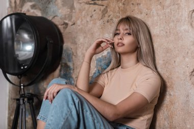 Sarı tişörtlü, mavi kot pantolonlu zarif, zarif genç bir bayan kapalı alandaki klasik tuğla bir duvarın yanında gevşemekten hoşlanıyor. Çekici kız modeli odada modern siyah bir lambanın yanında rahatlıyor..