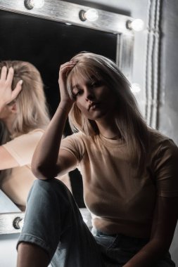 Modern, güzel, sarışın, şık bir saç stili olan, mavi kot pantolonlu, şık bir tişörtü olan, şık, beyaz bir aynanın yanında oturan, parlak lambalı, güzel bir kadın. Karanlık bir odada güzel poz veren bir kız..