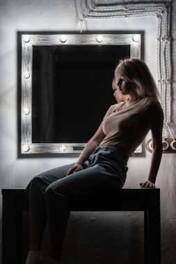 Şık, mavi kotlu, şık bir tişörtü olan genç, modaya uygun, sarışın bir kadın klasik beyaz bir aynanın yanında, parlak ampullü bir masanın üzerinde oturuyor. Akşam vakti evde seksi bir kız..