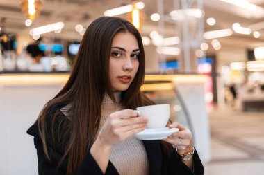 Modayı takip eden esmer, şık elbiseli bir kadının portresi kafede eski model bardakla. Zarif kız model içeride kahve molası vermeyi seviyor. Kafein severler için sabah.