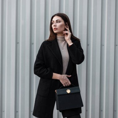Uzun saçlı, lüks moda siyah ceketli, deri çantalı, caddede metal duvarın yanında poz veren zarif, şık bir model genç kadın. Güzel kız modeli. Sevimli modacı bayan..