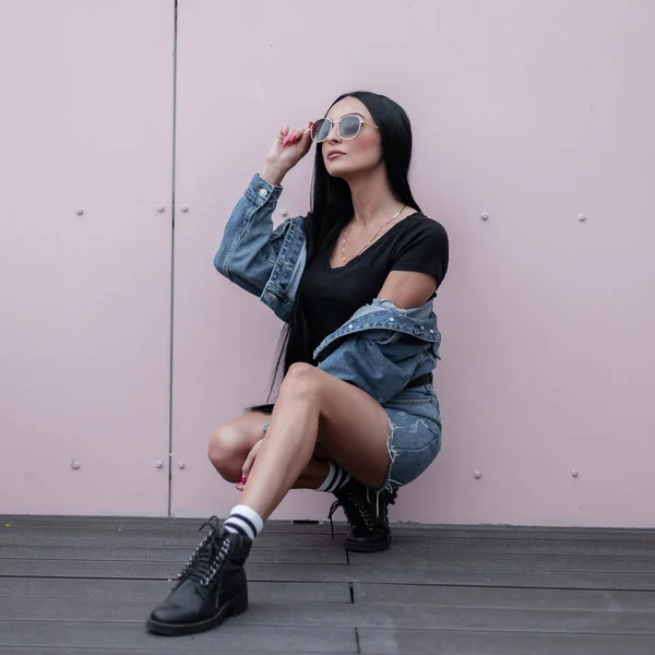 Schone Hubsche Junge Brunette Mit Sonnenbrille In Einem Trendigen Vintage Blauen Rock In Einem Stylischen Schwarzen Top In Einer Jeansjacke In Lederstiefeln Sitzt Neben Einer Wand Auf Einer Aussenterrasse Modisches Hipster Madchen Saison