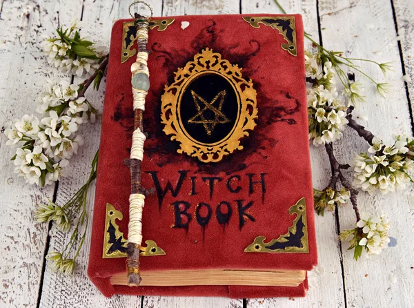 Witch book in red cover with pentagram, magic wand and spring flowers on table. Occult, esoteric and divination still life. Halloween background with vintage objects